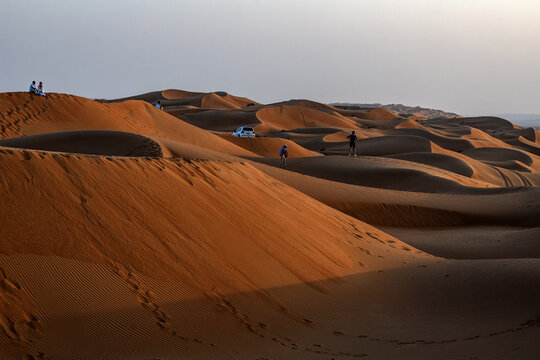 View of the vast sand dunes of Bidiyah with people walking across the undulating landscape under a soft golden light in Bidiyah, Ash Sharqiyah North G, Oman.