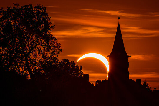 Partial solar eclipse above a church steeple and tree silhouettes against a dramatic orange evening sky, creating a striking celestial and spiritual scene.