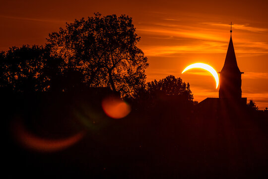 Partial solar eclipse above a church silhouette at sunset, with dramatic orange sky, dark trees and lens flare creating a mysterious atmospheric scene.