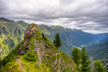 Obraz premium Hikers travel along a rocky path in the Ankogel Group. Lush green hills rise on either side. Clouds cover the sky. The scenery shows the beauty of nature in the Eastern Alps.