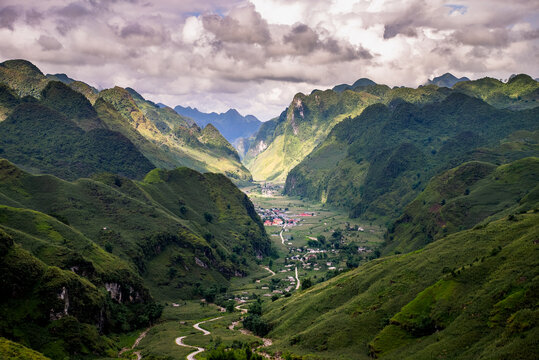 View of the Ha Giang Loop winding through lush green mountains and valleys with a small village nestled in the landscape under a cloudy sky in Dong Van, Ha Giang, Vietnam.