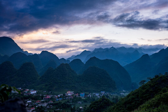 View of the dramatic karst mountain landscape of the Ha Giang Loop with a small village nestled in the valley under a cloudy sunset sky in Dong Van, Ha Giang, Vietnam.