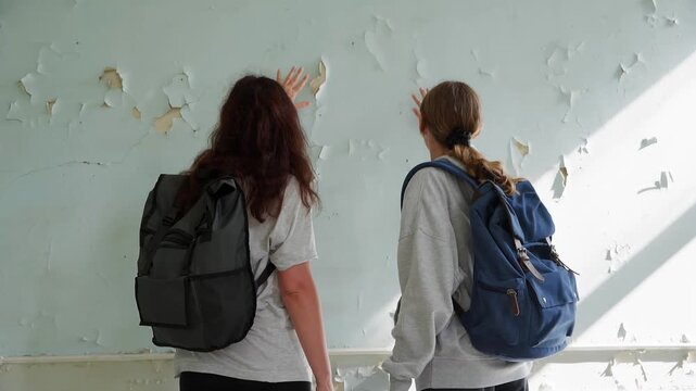 Two young women wearing backpacks stand facing crumbling plaster wall, one making expressive hand gesture toward peeling surface in sunlit room.