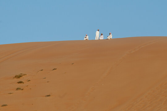 View of four men in traditional Omani clothing sitting on the crest of a sand dune under a clear blue sky in Bidiyah, Ash Sharqiyah North, Oman.