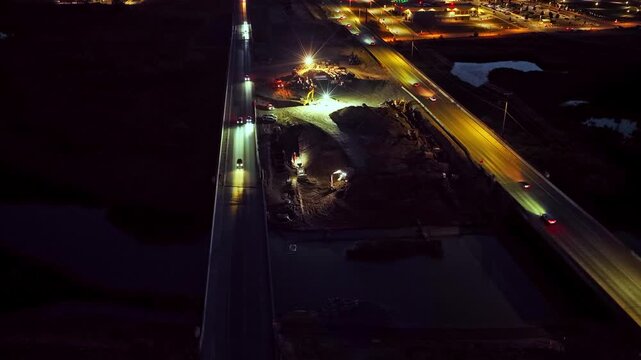 2100 North Freeway construction site in Lehi, Utah at night, with bright work lights illuminating heavy machinery, new roads, and traffic streaming by - static, wide angle, drone view