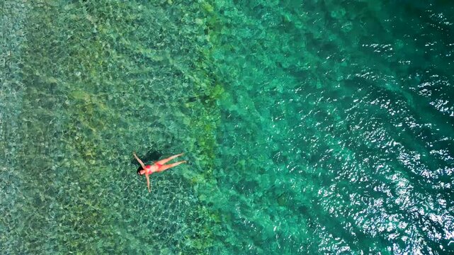 Woman in a red swimsuit swims on her back in the clear sea, top down view