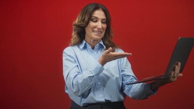 Woman holds laptop and presents palm up toward the screen against a red studio backdrop, smiling and gesturing an invitation; confidence approachability.