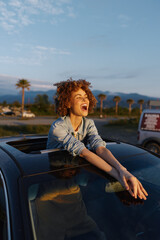 Obraz premium Candid lifestyle portrait of a joyful woman leaning from a car window, outdoors at golden hour, palm trees in the background, capturing carefree travel mood and radiant happiness