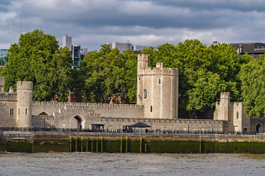 View of the historic stone walls and towers of the Tower of London, including Traitors' Gate, framed by lush green trees under a cloudy sky in London, England, United Kingdom.