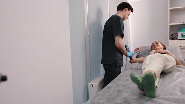 Vibration massager, Handheld massager, Percussion device. Chiropractor, in dark scrubs and gloves, uses percussion massager on arm of female patient lying on treatment table.
