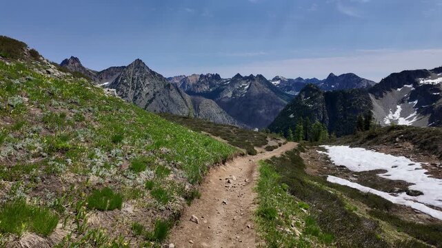 First-person point of view moving forward along a dirt mountain path with snowfield patches and dramatic mountain range in the background on a sunny day in the North Cascades, Washington