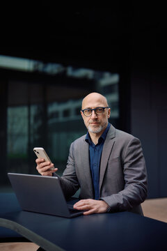 Businessman using cell phone and laptop in front of office building and looking at camera.