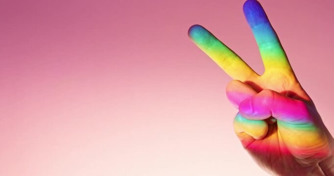 LGBTQ+ Pride Victory and Peace Gesture in Rainbow Light over Pink Background in slow motion with copy space. Symbolic LGBTQ+ Pride concept representing love, equality, diversity, inclusion, acceptance