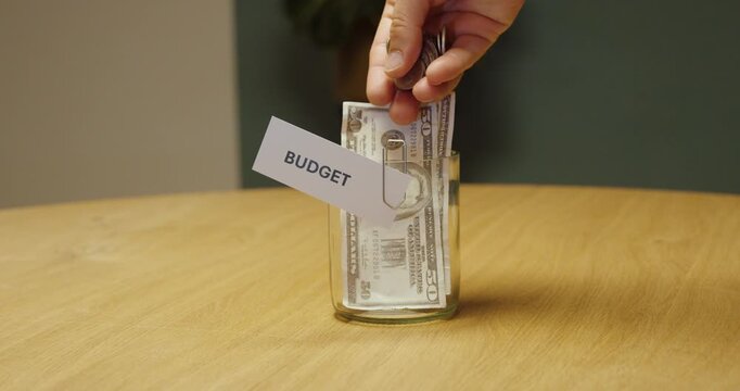 Hand placing a few coins into a jar labeled &ldquo;Budget,&rdquo; representing a small budget, limited financial resources, cost constraints and careful money management.