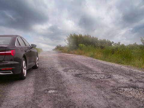 Black car on rough, potholed road. Misty weather, overcast sky. Green vegetation lines the roadside. Road stretches into distance, blurred by fog. Living in remote country side with bad road surface