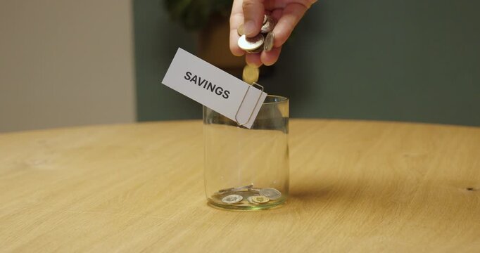 Hand placing coins into a glass jar labeled &ldquo;Savings,&rdquo; representing saving money, personal finance, budgeting and financial planning.