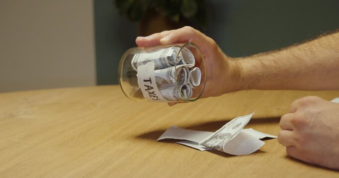 Person emptying money from a jar labeled &ldquo;Taxes,&rdquo; representing tax payments, financial burden, rising expenses and personal budgeting.
