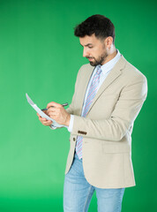 Adult man stands against green screen, holding paper documents and writing notes or filling forms with pen in hand © JackF