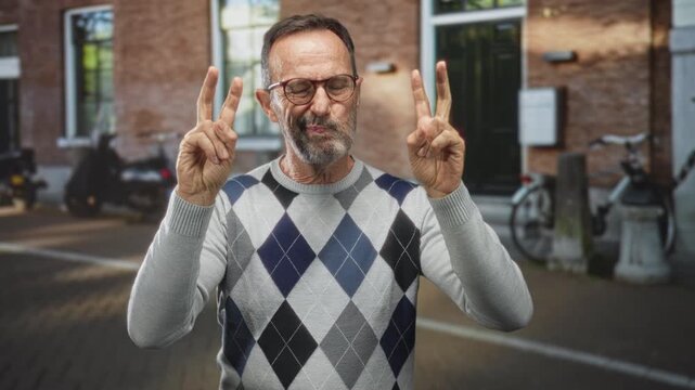 Man, middle aged, wearing argyle sweater and glasses, shows peace signs with both hands on a street beside a brick building; playful confidence.
