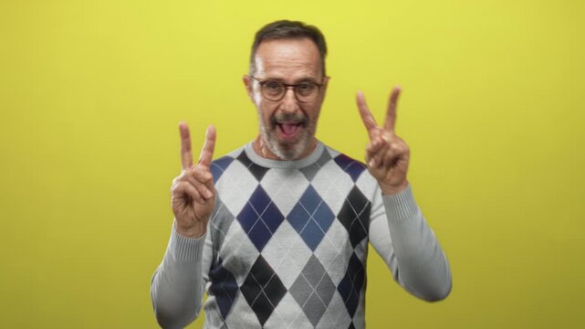 Man with glasses and argyle sweater holds up peace signs with both hands in a yellow studio; playful celebration.