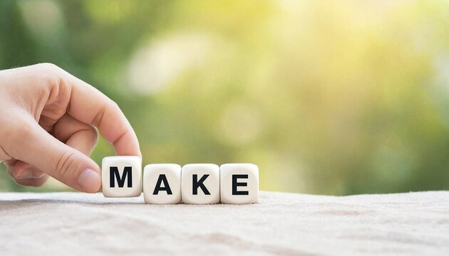 Hand arranging white letter cubes spelling the word make on a soft fabric surface with green bokeh background