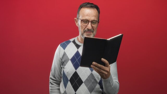 Man reading and holding a black book with hand in studio against red backdrop, wearing glasses and argyle sweater; contemplation.