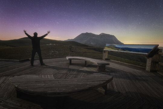 View of a person standing on a wooden observation deck at Forca di Presta with arms raised towards the starry night sky and mountain landscape in Arquata del Tronto, Marche, Italy.