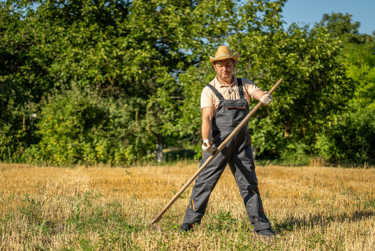 A gardener stands in a dry field holding a long tool. He wears a straw hat and overalls. Green trees surround the area. It is bright and sunny