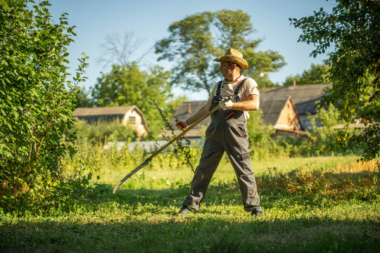 A farmer stands in a green field using a scythe to cut grass. In the background, there are farm buildings and trees under a clear sky. It is daytime and bright outside