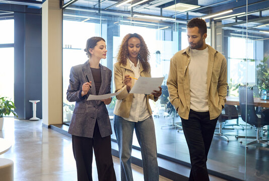 Colleagues in office discussion review business documents. Three professionals walk along a glass corridor, comparing pages and aligning next steps. Clear theme of teamwork and planning.