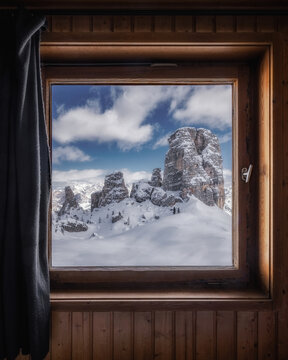 View of the snow-covered Cinque Torri rock formations seen through a rustic wooden window frame in the Dolomites, Cortina d'Ampezzo, Veneto, Italy.