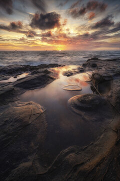 View of the rocky coastline at sunset with tide pools reflecting the dramatic clouds and golden light in Calafuria, Toscana, Italy.