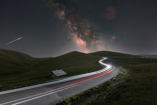 View of the Milky Way and light trails on a winding road at Campo Imperatore with a small stone hut nestled in the grassy hills of L'Aquila, Abruzzo, Italy.