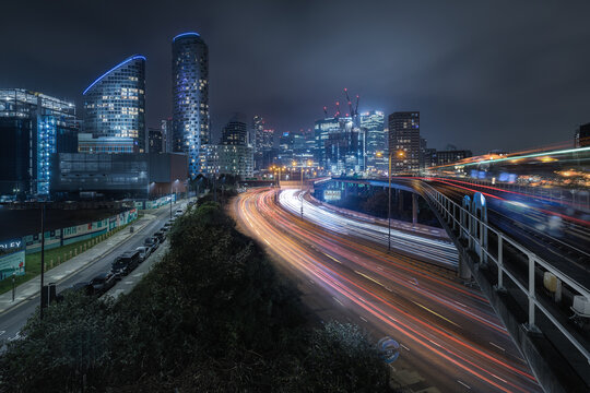 View of the illuminated Canary Wharf skyline at night with light trails from traffic and the DLR train tracks in London, England, United Kingdom.