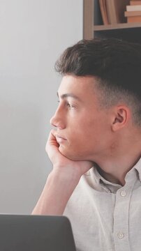 Portrait of a pensive and tired young man looking away from his laptop screen, feeling bored and unmotivated while studying or working on a difficult task in his home office or library