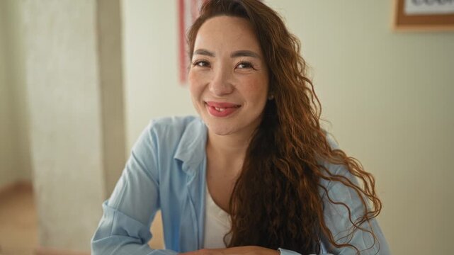 Woman smiles at camera with hands resting on desk, long curly hair and usa flag on wall in a building office; calm confidence.