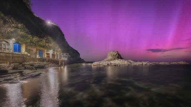 View of the Chair of the Pope rock formation illuminated by a vibrant purple aurora borealis above the rocky coastline and grottos of Ancona, Marche, Italy.