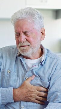 Elderly man with white hair feeling intense chest pain, holding his heart and showing a painful expression, potentially experiencing a myocardial infarction or heart attack at home