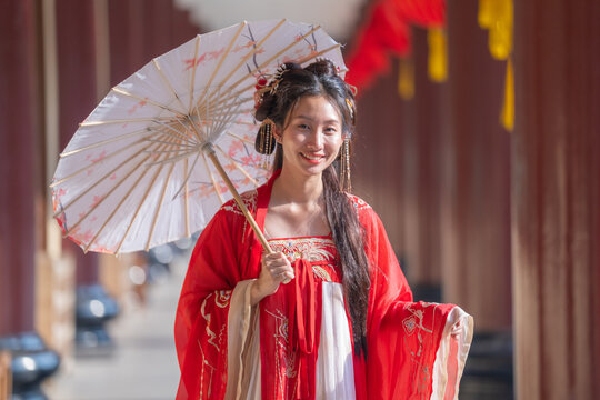 A young Chinese woman wearing an embroidered red hanfu robe, smiles brightly, parasol angled overhead, at red pillared corridor, round red lanterns with yellow tassels, potted greenery.