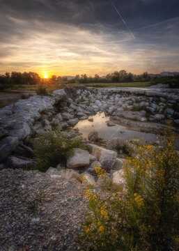 View of the rocky riverbed of the Lungo Tenna at sunset, with yellow wildflowers in the foreground and golden hour light reflecting on the water, Lungo Tenna, Marche, Italy.