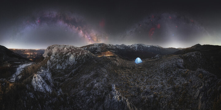 View of a small blue tent pitched on a rocky mountain ridge under the vibrant arc of the Milky Way galaxy in the night sky in Bolognola, Marche, Italy.
