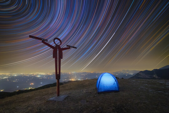 View of the Punto panoramico con croce sculpture and a glowing blue tent on a mountain top under a night sky with vibrant star trails and distant city lights in Sarnano, Marche, Italy.