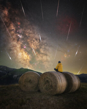 View of a person in a yellow jacket sitting on a hay bale at night, observing a meteor shower and the Milky Way galaxy above the landscape of Sassotetto, Marche, Italy.