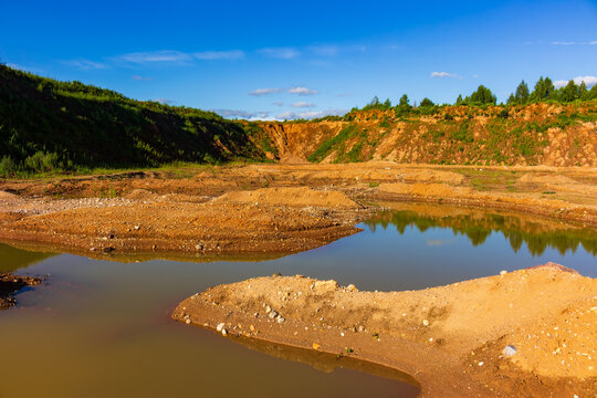 Exposed sandy soil terrain featuring small water pools after industrial sand extraction, surrounded by steep weathered slopes under a bright blue summer sky in an abandoned quarry