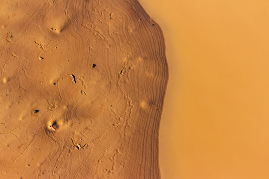 Close-up of a muddy clay puddle with textured sandy banks at a quarry, creating a surreal alien landscape with ripples and abstract earthy patterns in orange and brown tones