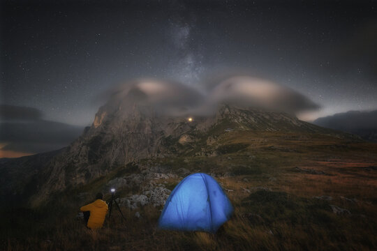 View of a camper in a yellow jacket sitting next to a blue tent under a starry sky with the Milky Way above the lenticular clouds covering the peaks of Prati di Tivo, Abruzzo, Italy.
