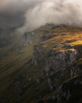 View of the dramatic rocky cliffs and grassy mountain slopes illuminated by golden sunlight under a blanket of clouds in Prati di Tivo, Abruzzo, Italy.