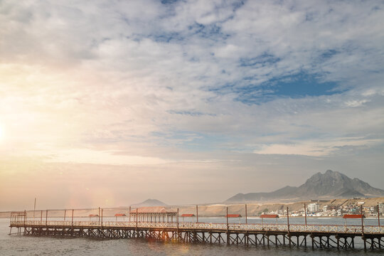 Panoramic view of Huanchaco wooden pier extending over Pacific Ocean with dramatic sunset sky and mountain peak in background