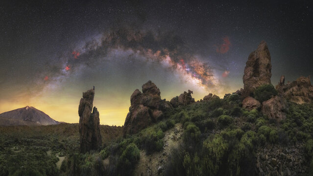View of the iconic Roque Cinchado rock formation under the vibrant Milky Way galaxy with Mount Teide in the background in Santa Cruz de Tenerife, Canary Islands, Spain.