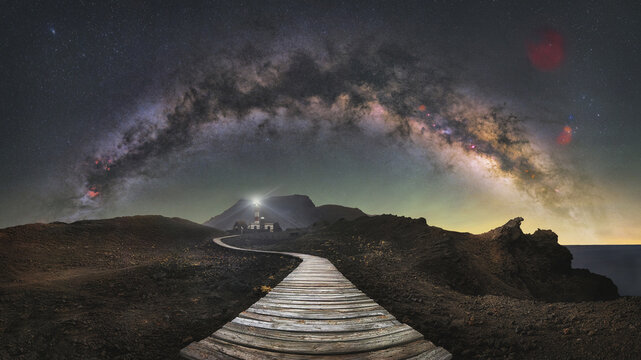 View of Punta Teno Lighthouse at night with a wooden path leading towards it under the arch of the Milky Way galaxy in a volcanic landscape, Buenavista del Norte, Canarias, Spain.